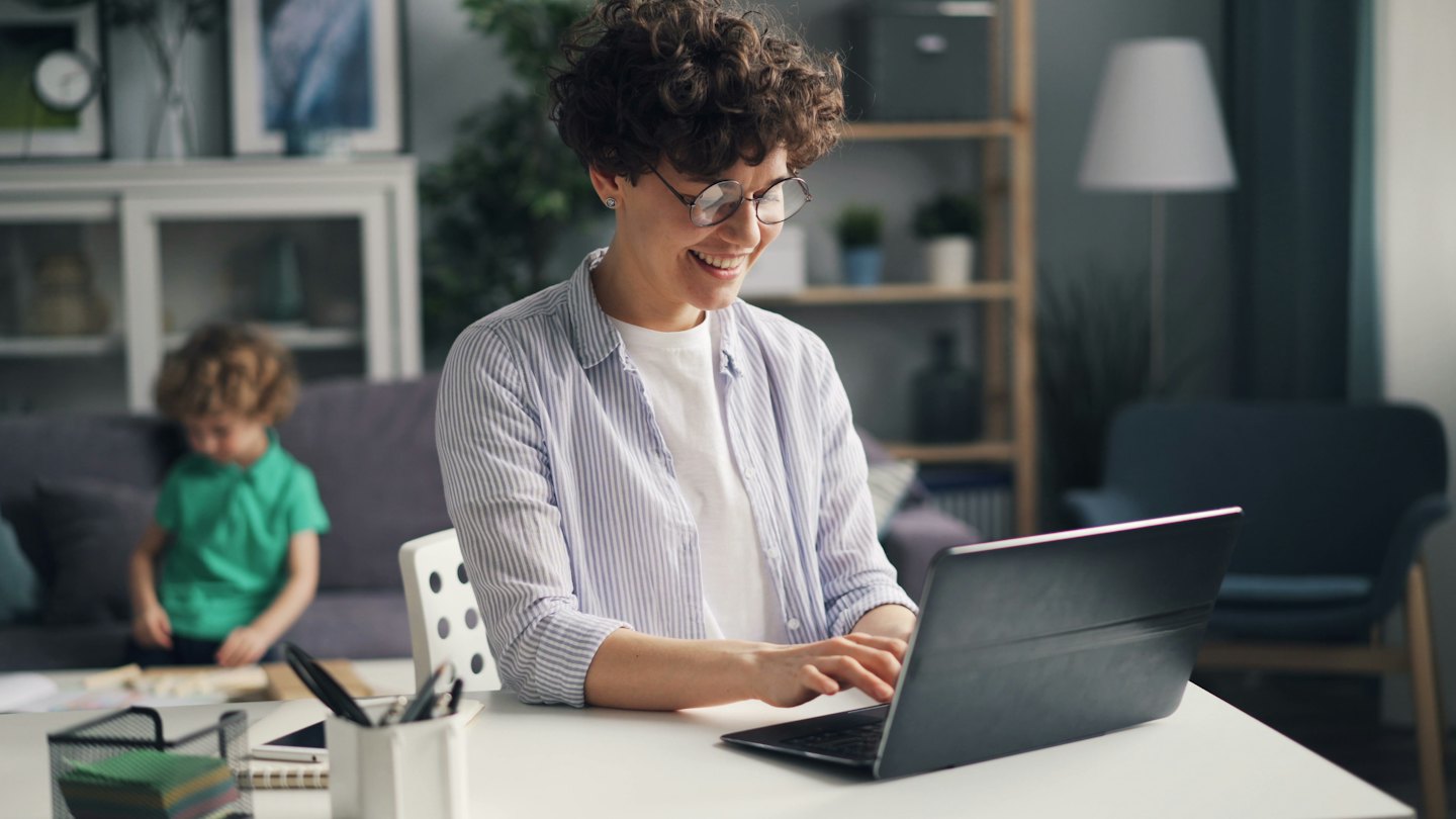 Eine Frau lächelt, während sie am Schreibtisch auf einem Laptop tippt, mit einem Kind im Hintergrund.